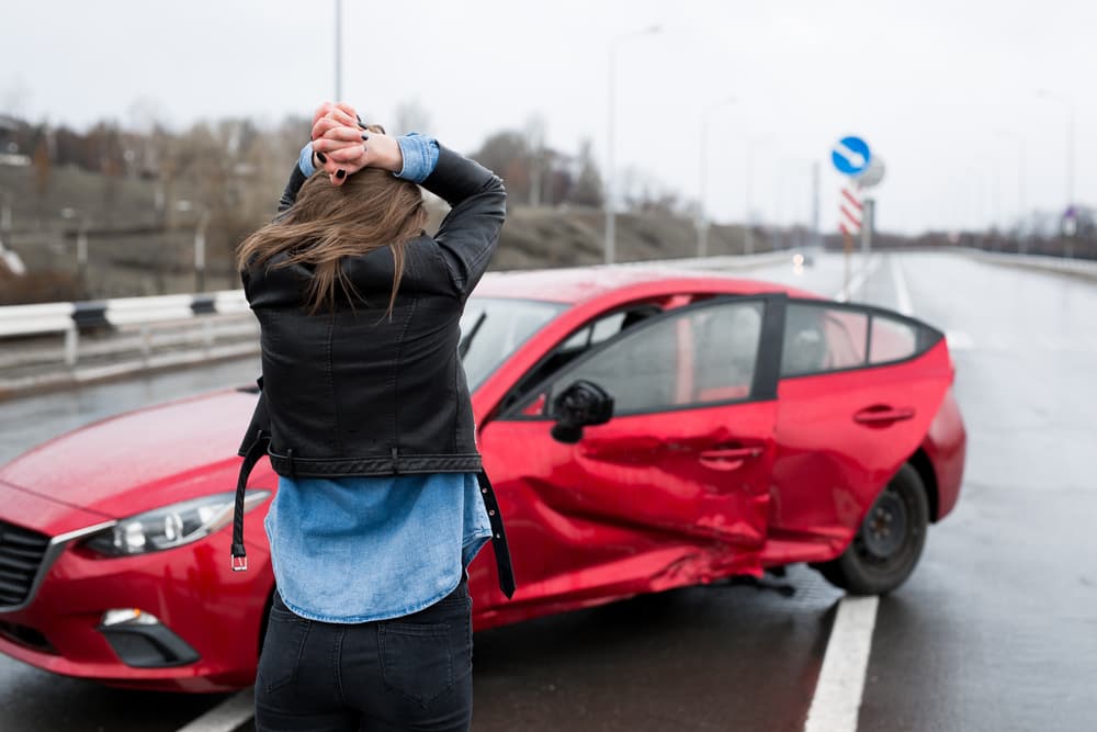 Woman stands beside a damaged car after an accident, signaling for help and dealing with car insurance.