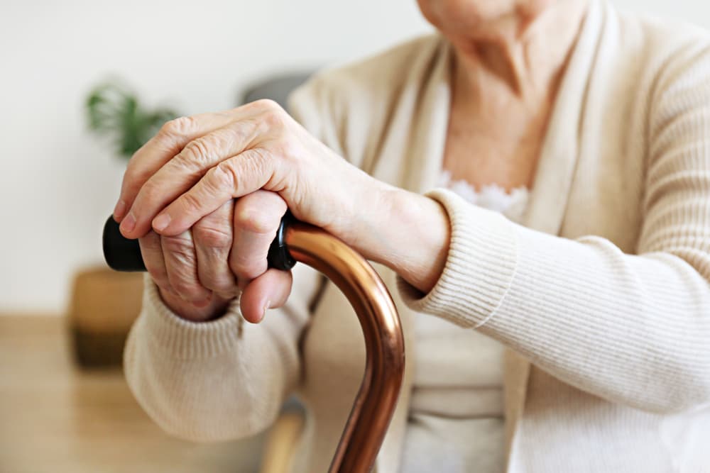 An elderly woman sits in her nursing home room, holding onto a quad cane for support. Her hands, weathered with age, gently grasp the metal handle.